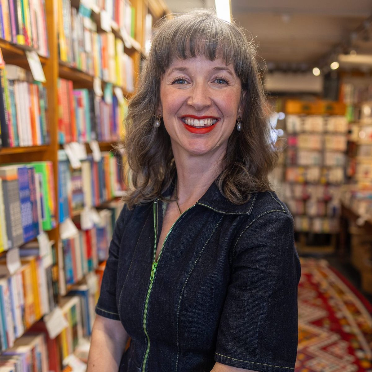 Woman smiling in a bookshop aisle, wearing a dark denim dress, with bookshelves lining the walls behind her.