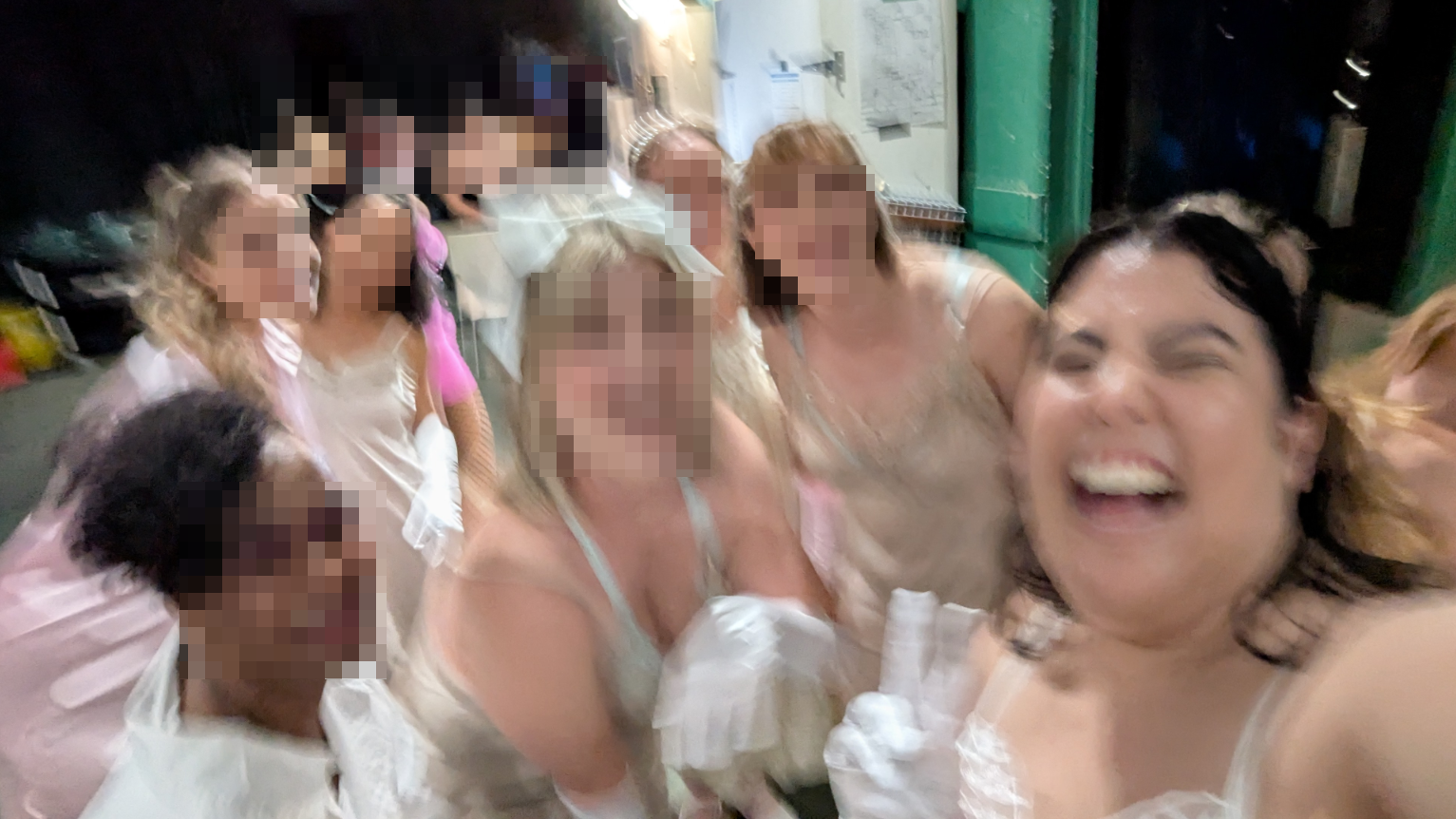 A joyful, slightly blurry backstage selfie of myself and a group of burlesque classmates in costume. Everyone is dressed in light-colored satin slips, gloves, and accessories, smiling and laughing together. Faces of everyone apart from myself are pixellated for their privacy.