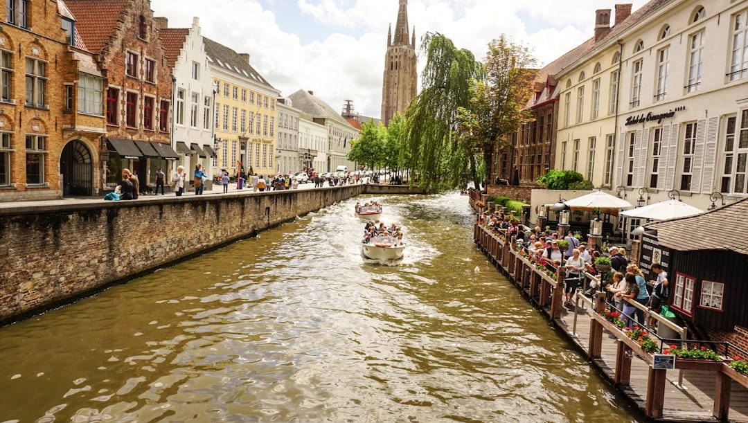 Open-topped tour boats carrying passengers along a Bruges canal lined with brick houses, the Church of Our Lady tower in the distance
