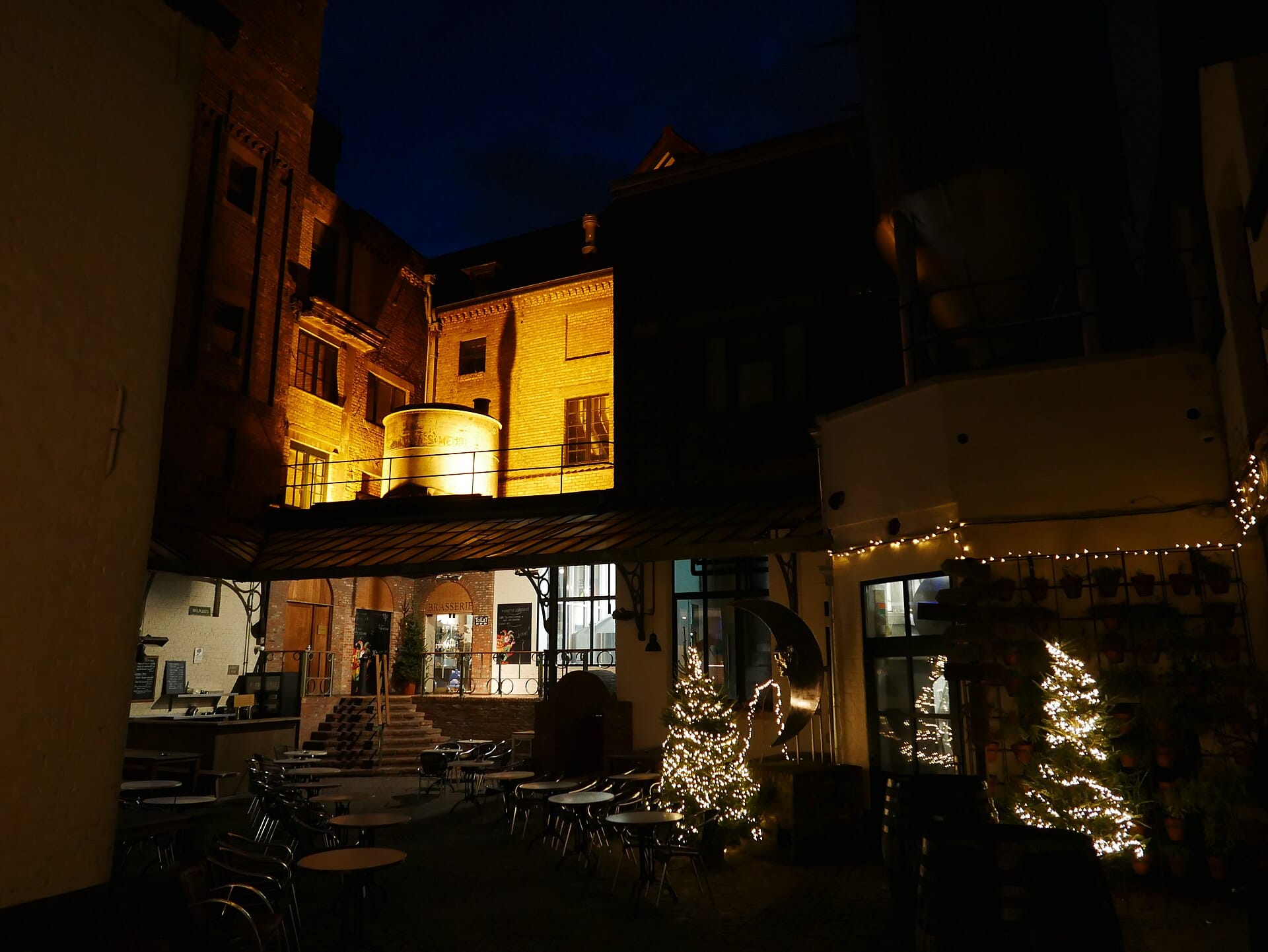 The lamplit courtyard of De Halve Maan brewery in Bruges at dusk, with its brasserie and a crescent-moon sculpture