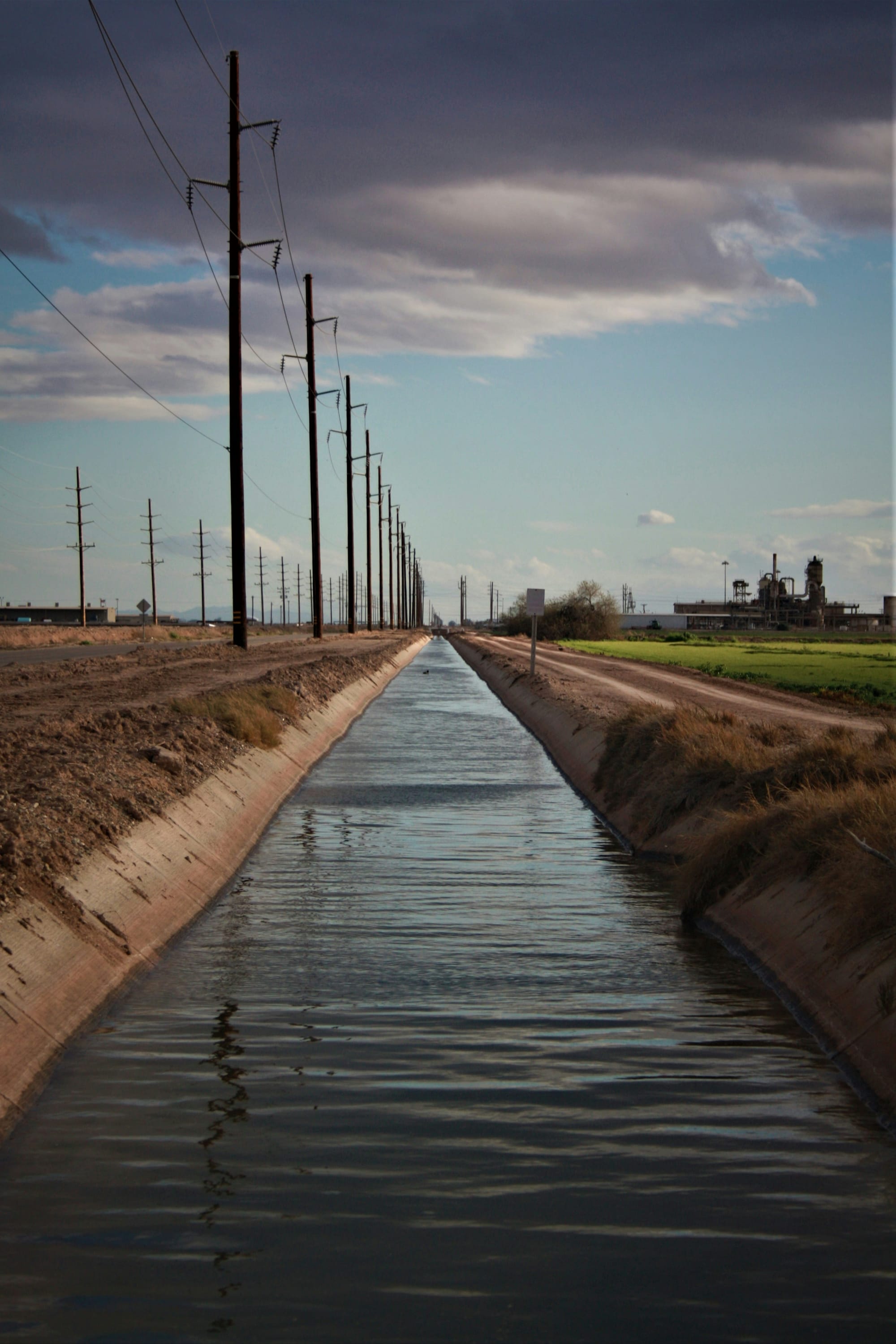 A concrete irrigation canal running through dry land beside power lines and an industrial plant.
