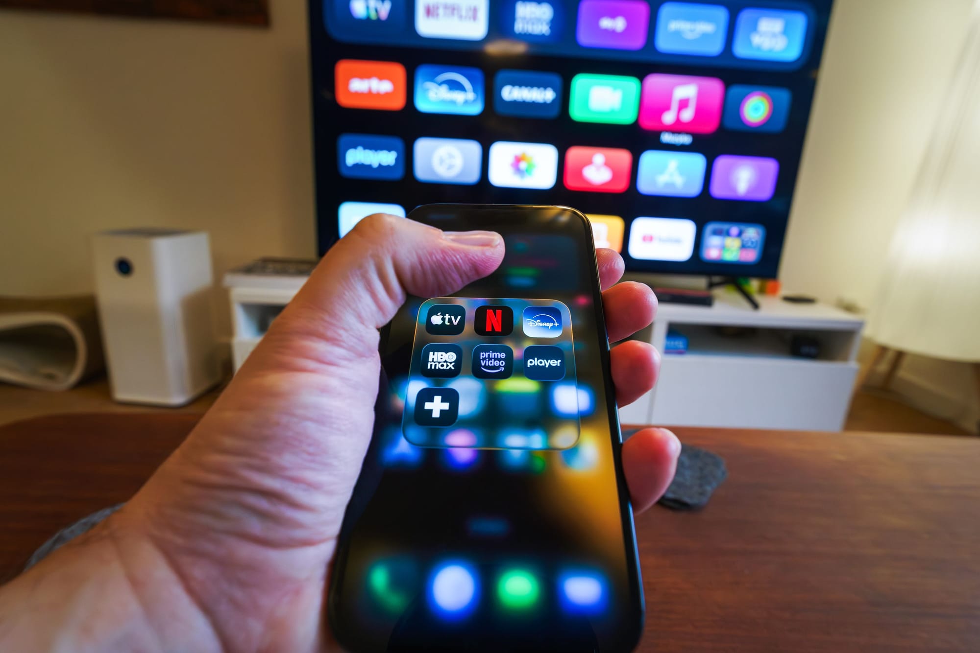 A hand holding a phone in front of a smart TV, both screens filled with rows of streaming and app icons.