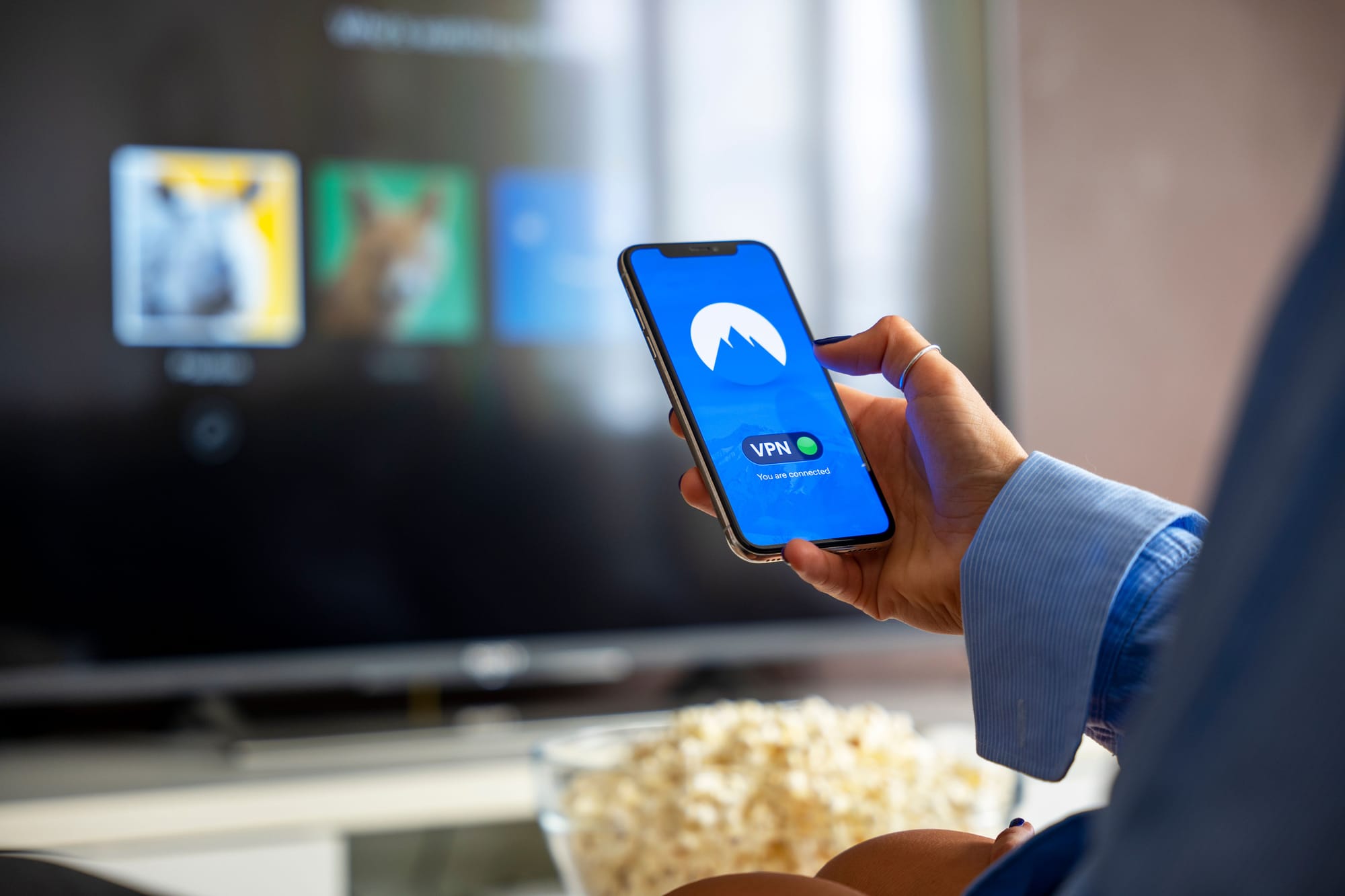 A hand holding a phone that displays a VPN app's "connected" screen, with a smart TV blurred in the background