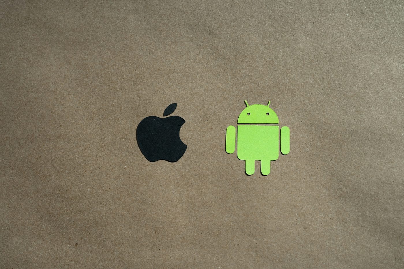 Paper cutouts of the Apple logo and the green Android robot side by side on a kraft-paper background.