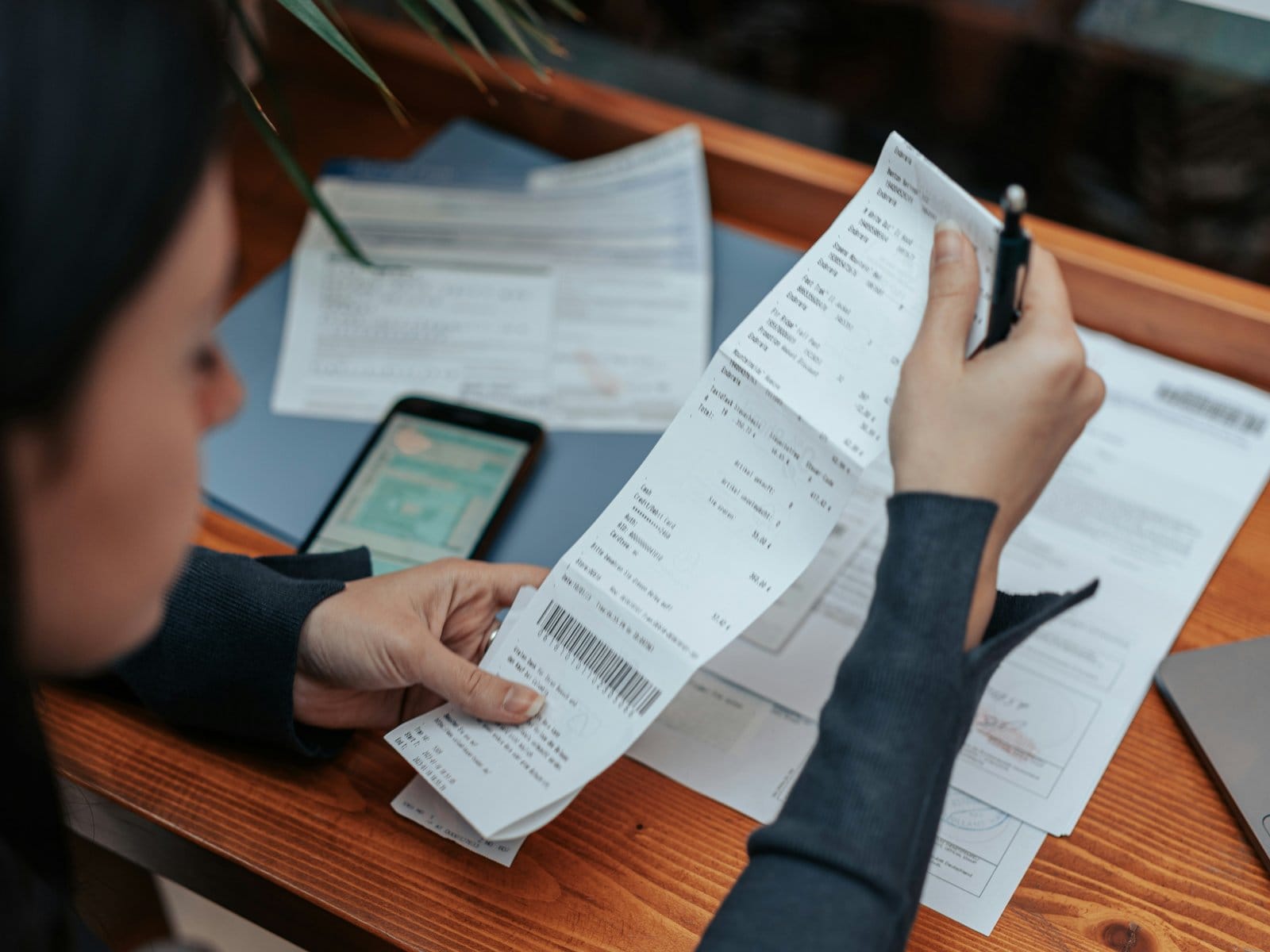 A person at a wooden table reviewing a long receipt, with bills and a phone in front of them