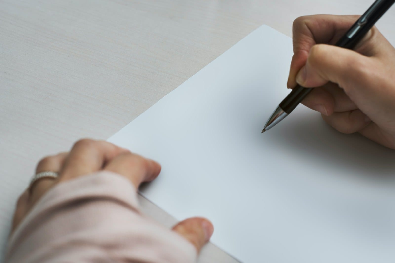Two hands at a table, one holding a pen over a blank sheet of paper.