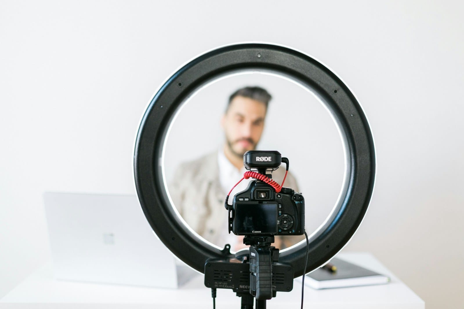 A camera and microphone mounted inside a ring light, aimed at a man sitting out of focus in the background