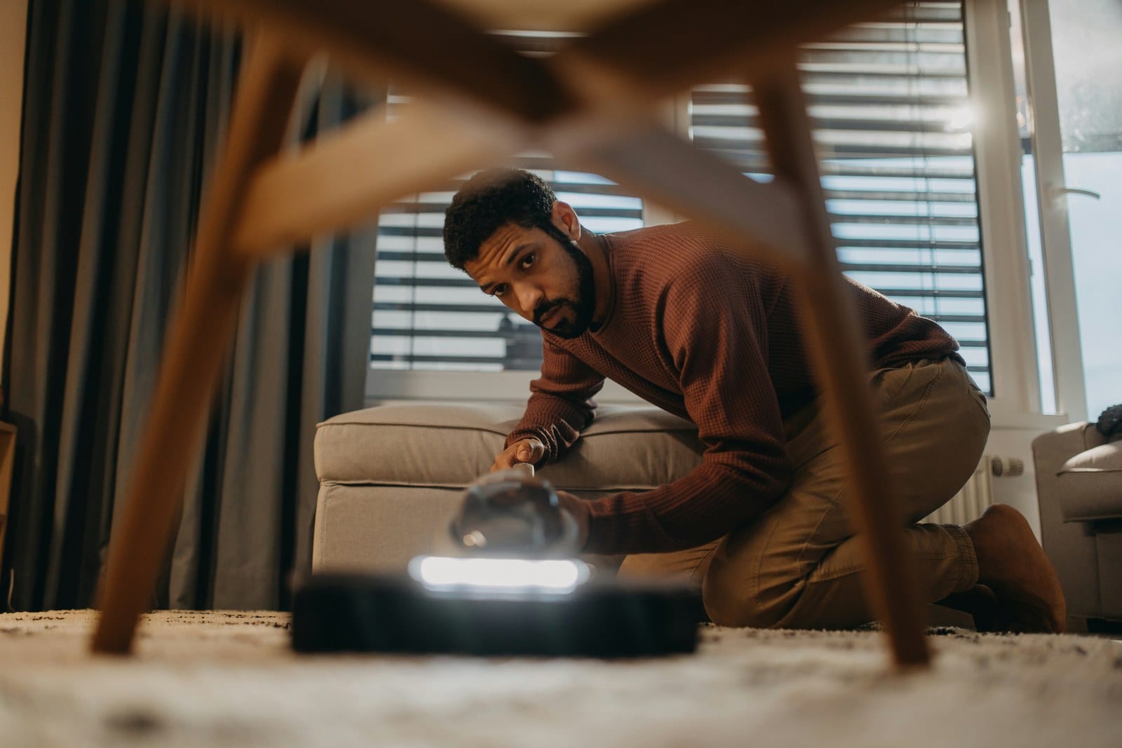 A man kneeling on a living room floor, looking under a table at a robot vacuum
