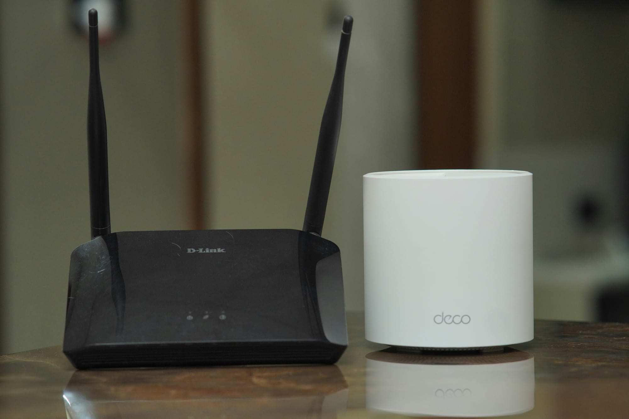 A black two-antenna D-Link router beside a white cylindrical TP-Link Deco mesh unit on a table.