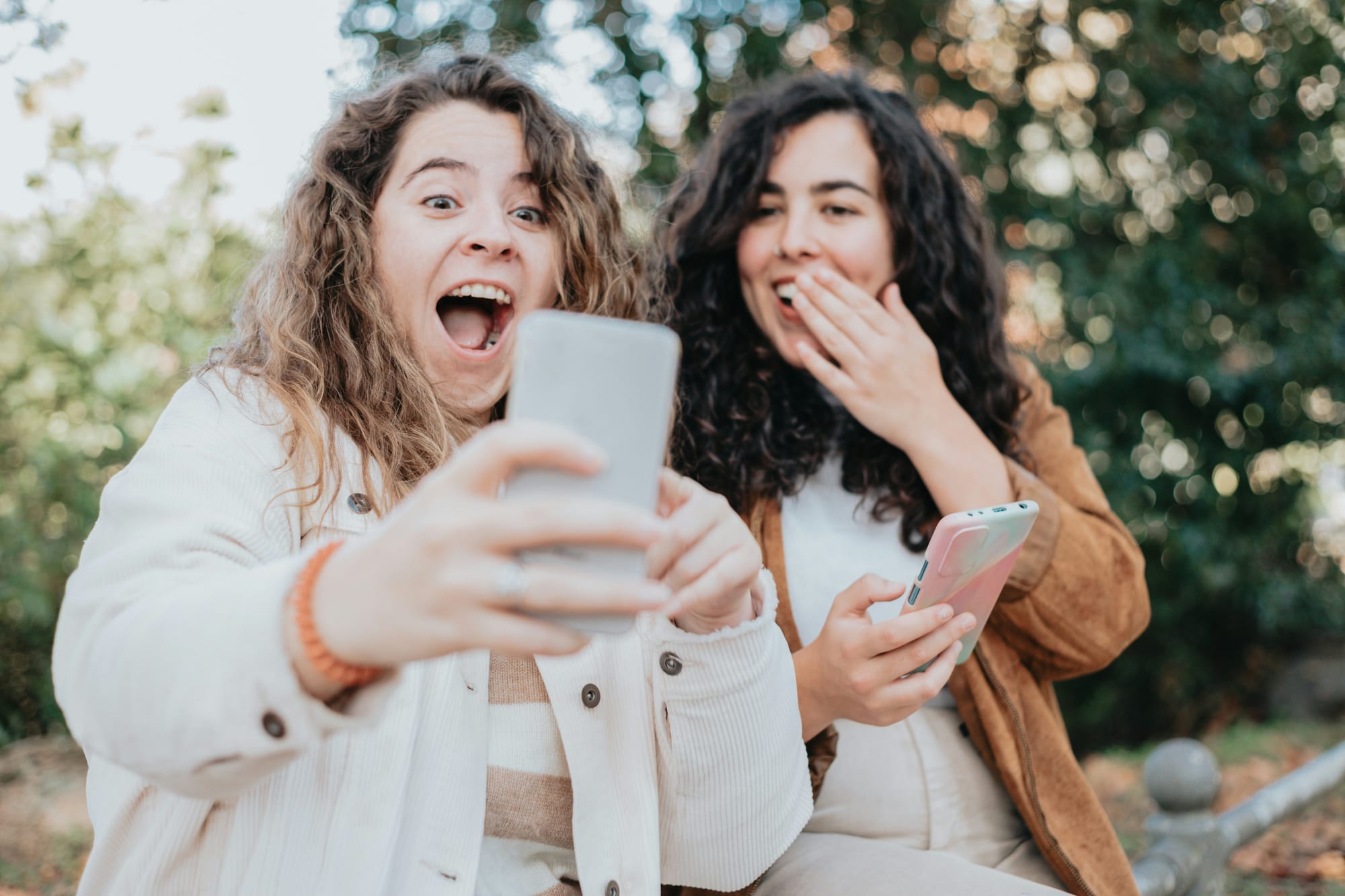 Two women sitting outdoors laughing, one holding up a phone with a delighted expression.