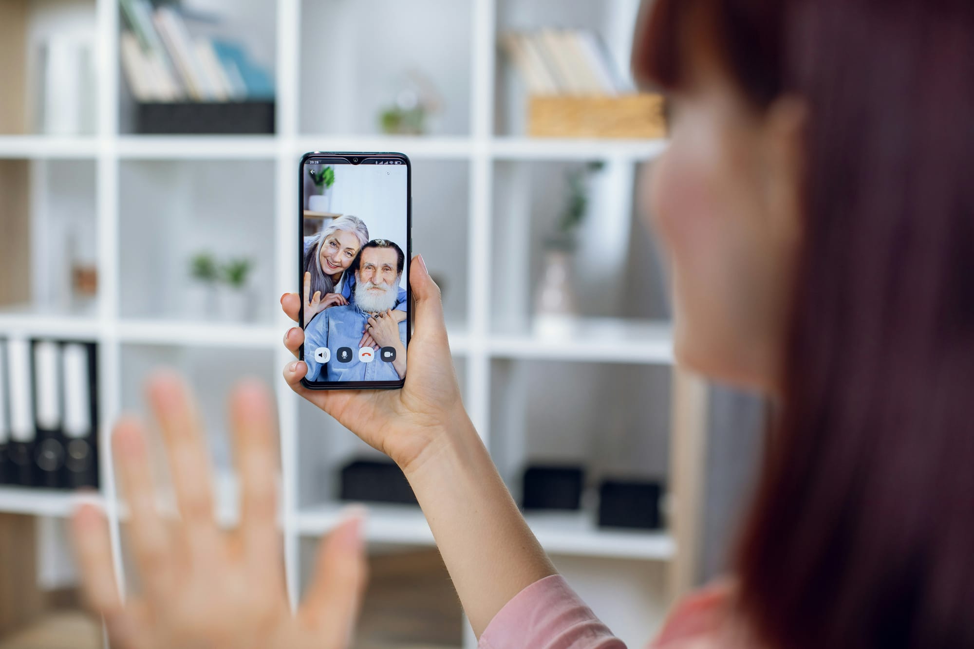 A woman holds up a phone showing an older couple waving back at her on a video call.