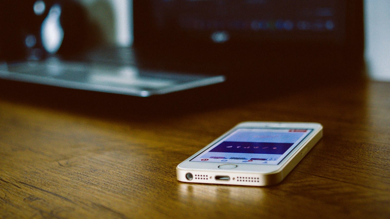 A smartphone lies face up on a wooden desk with an open laptop blurred behind it.