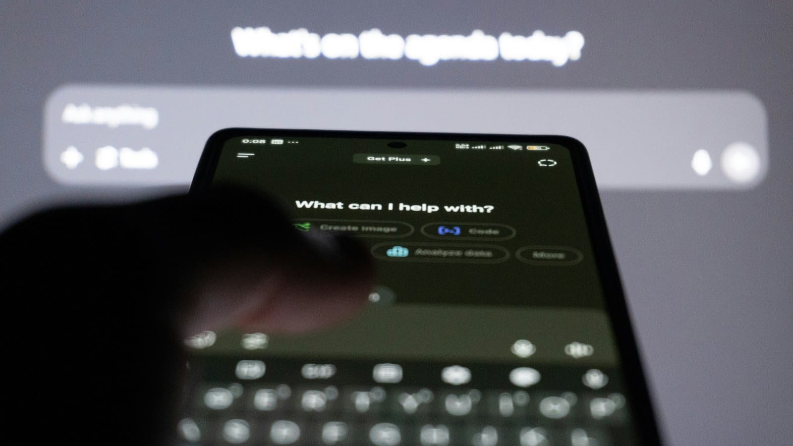 A hand holding a phone showing an AI assistant asking what it can help with, in front of a larger screen.
