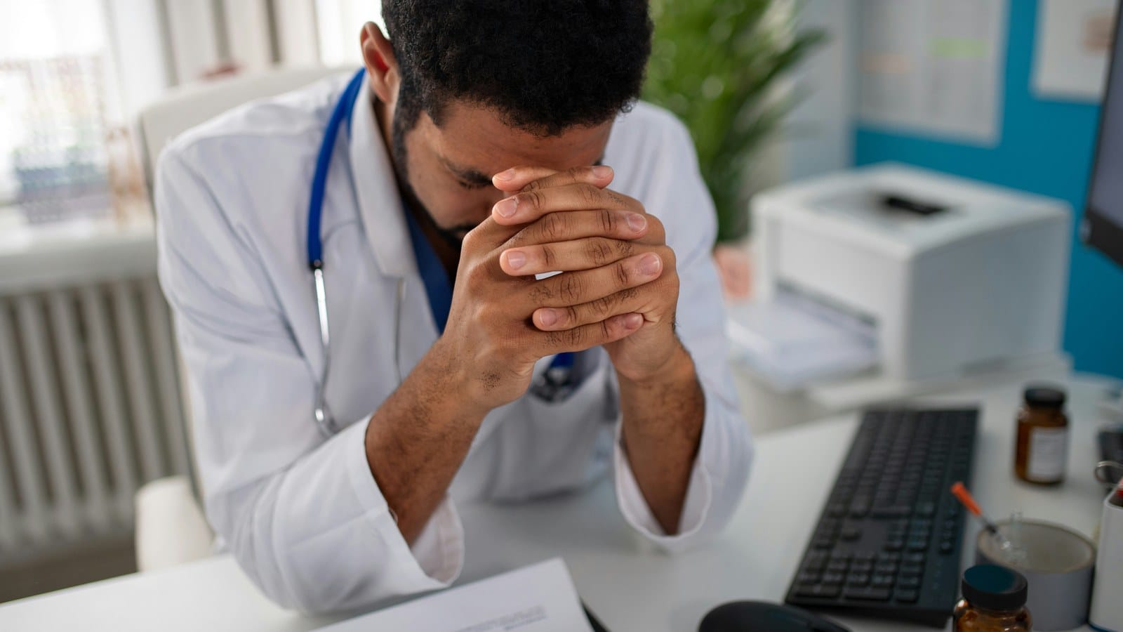 A doctor sits at a desk with his head bowed and hands clasped against his forehead, a keyboard and paperwork in front of him.