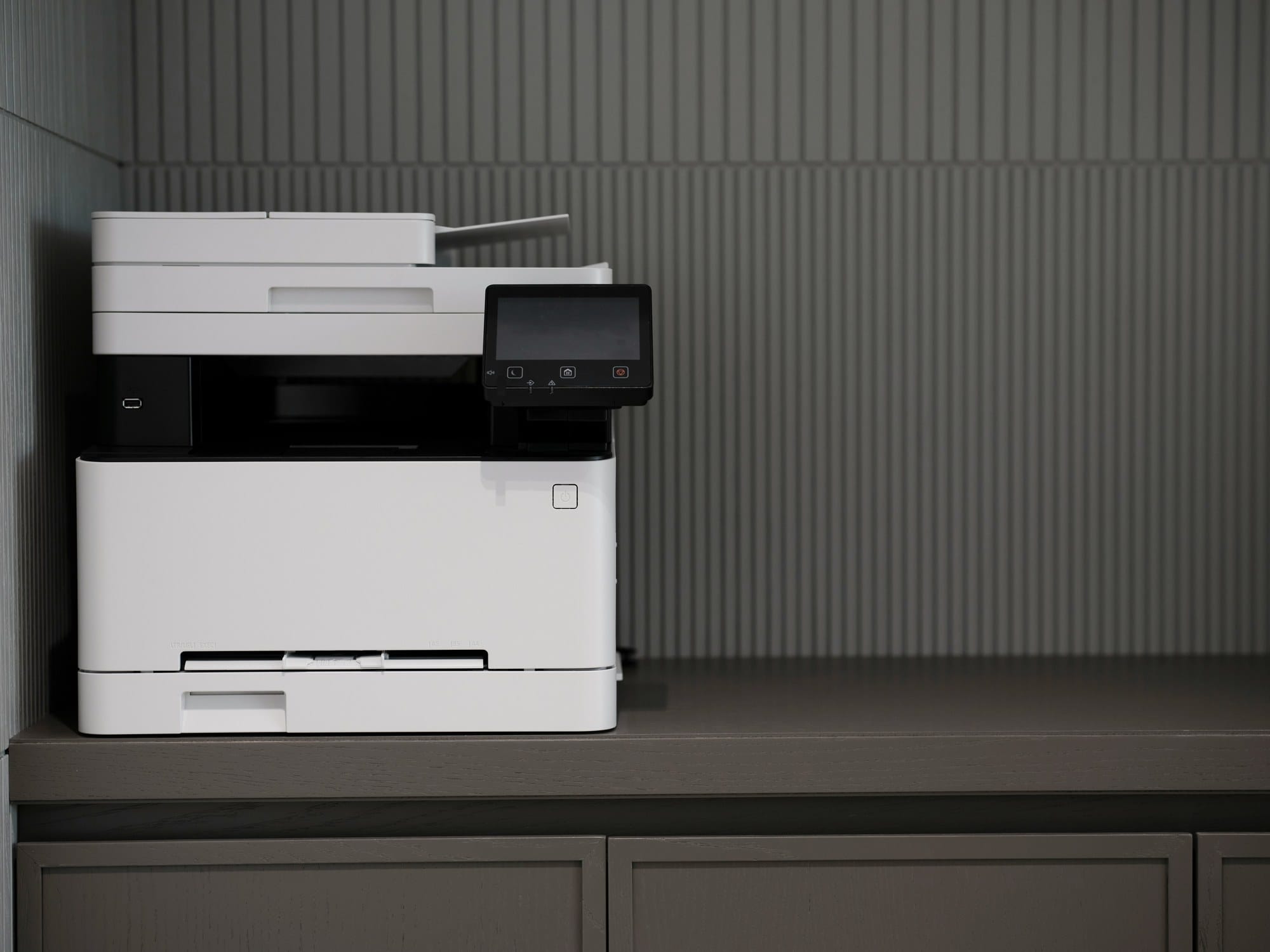 A white laser multifunction printer on a credenza against a grey fluted wall