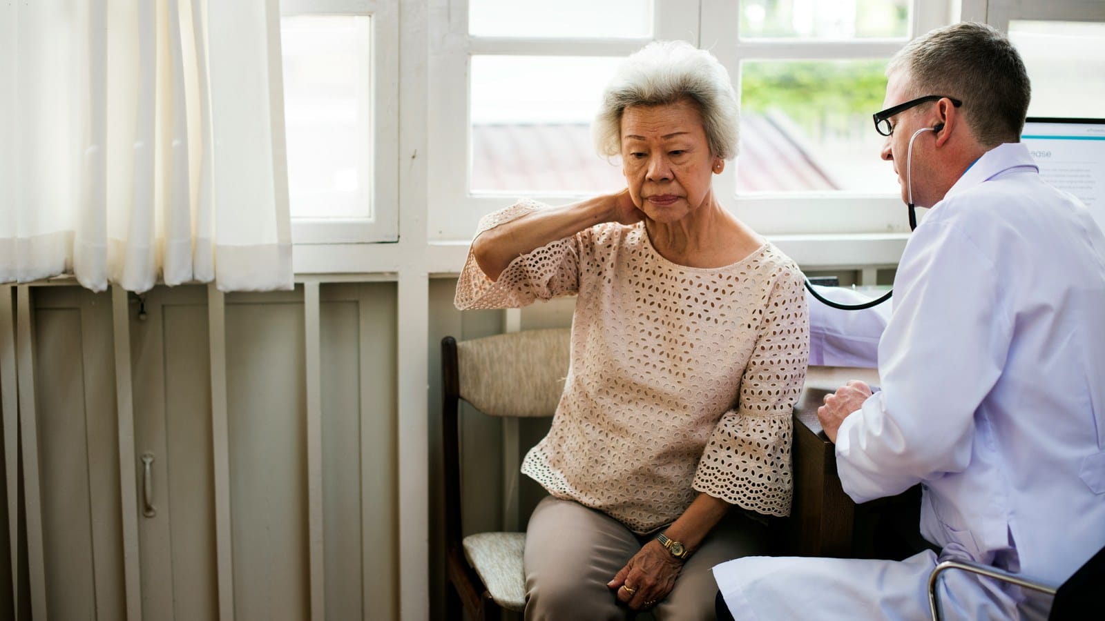 An older woman sits in a clinic chair holding the back of her neck while a doctor with a stethoscope listens beside her.