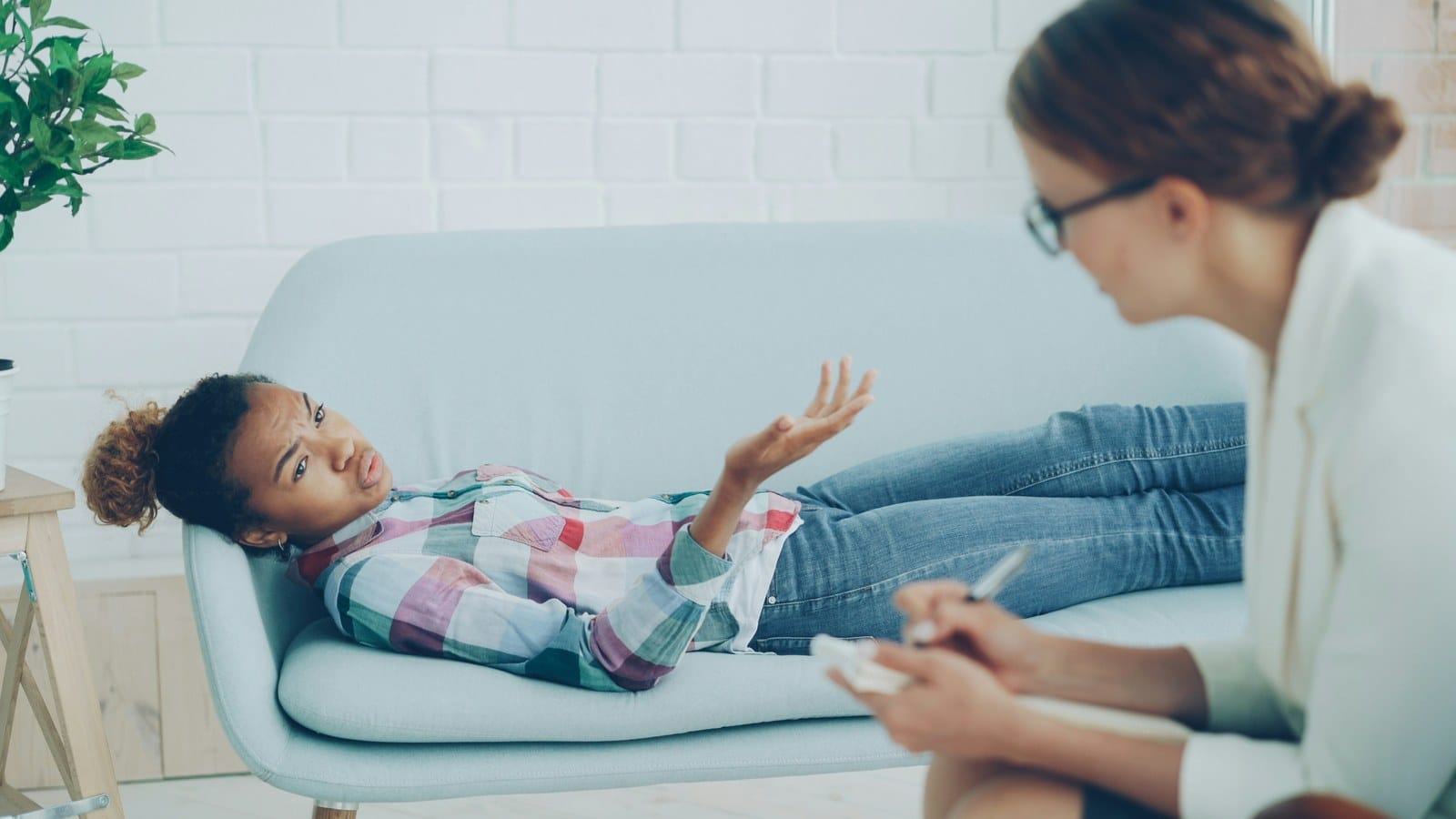 A young woman lies on a couch talking with her hands raised while a therapist takes notes on a pad in the foreground.