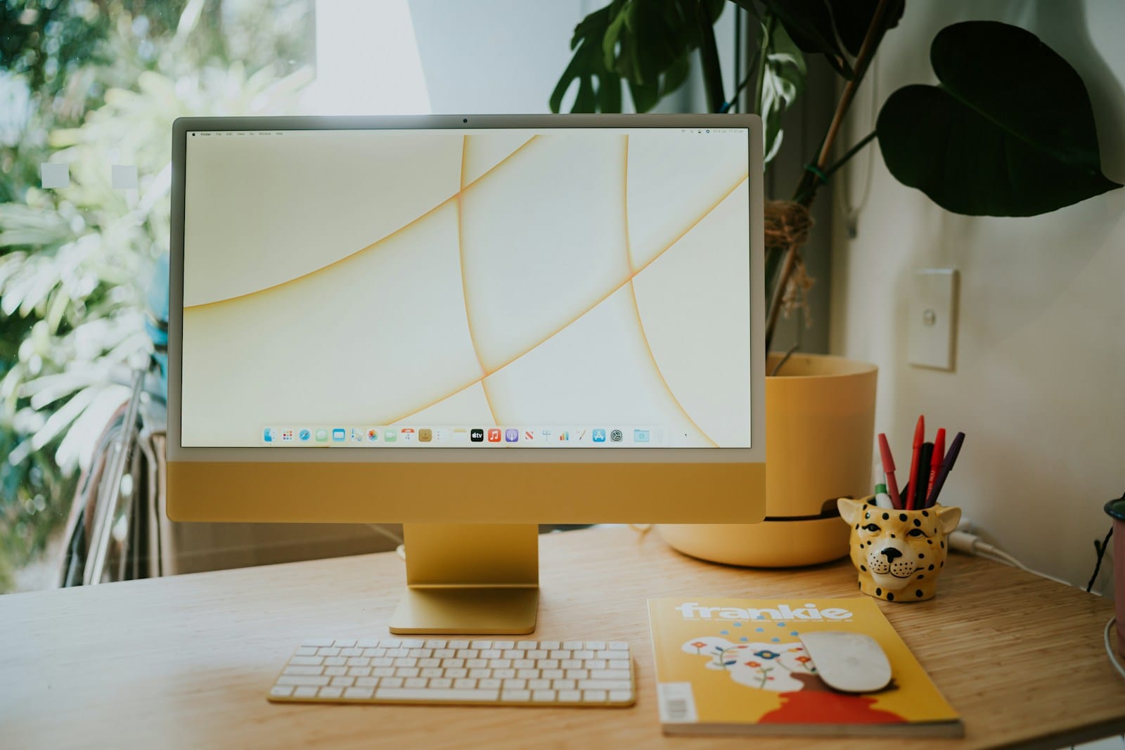A yellow iMac on a home desk beside a plant, a magazine, and a pot of pens