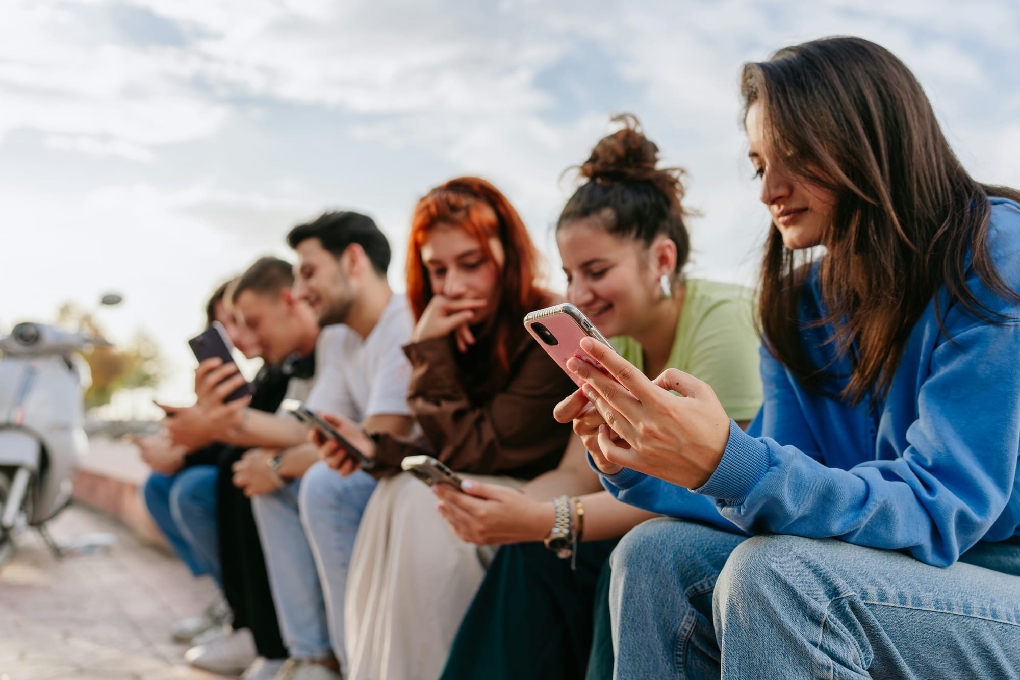 Six young people sit shoulder to shoulder along a low wall outdoors, each looking down at their own phone.