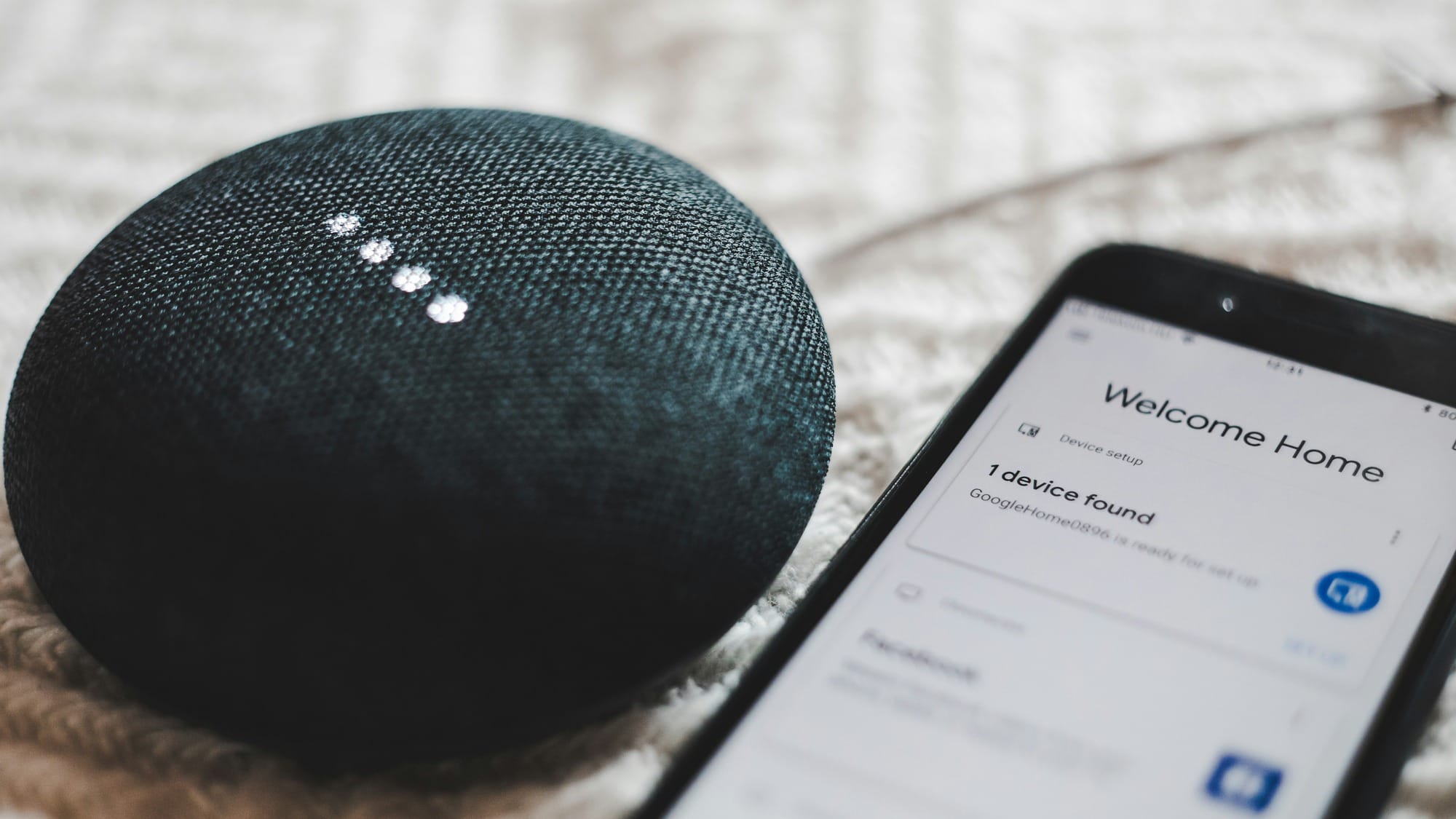 A small round fabric-covered smart speaker on a blanket, its four status lights lit, beside a phone showing a device setup screen that reads Welcome Home