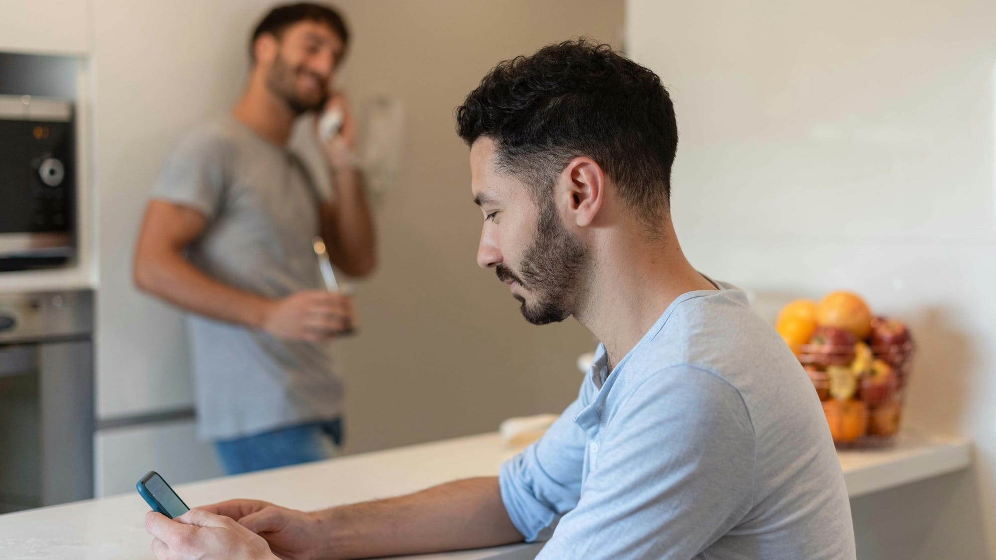 Two men in a kitchen. One sits at the counter looking at the phone in his hands while the other stands behind him talking on a phone