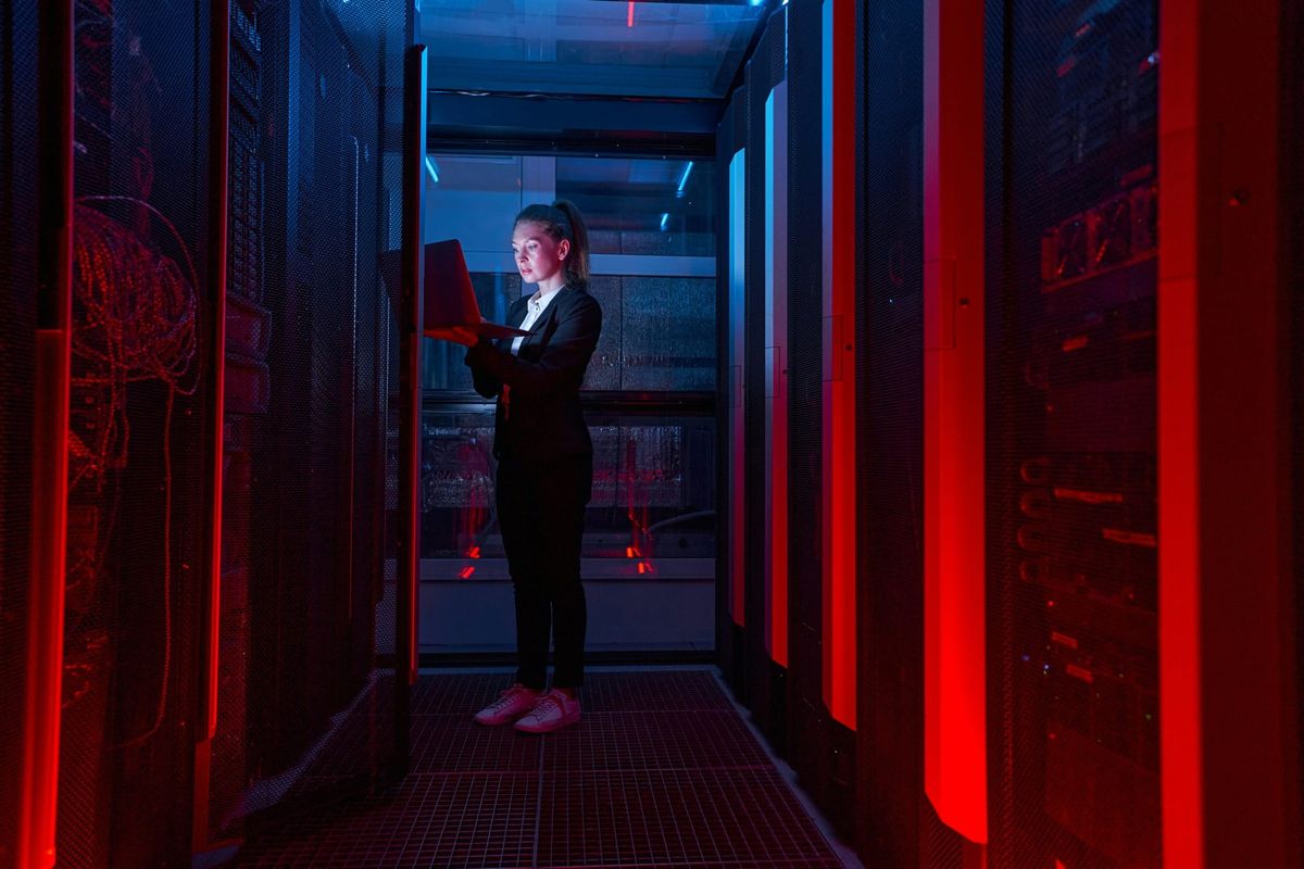 A security analyst works on a laptop in a dark, red-lit server room.