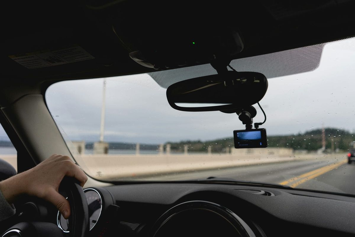 The view from a car's driver seat: a hand on the wheel, a small recording device by the mirror, an open bridge road ahead.