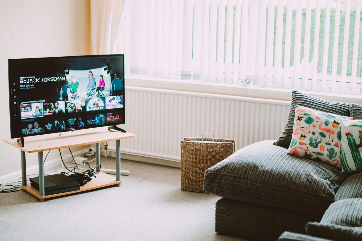 A flat-screen smart TV on a low stand in a bright living room, showing a streaming app's home screen, with an empty couch beside it