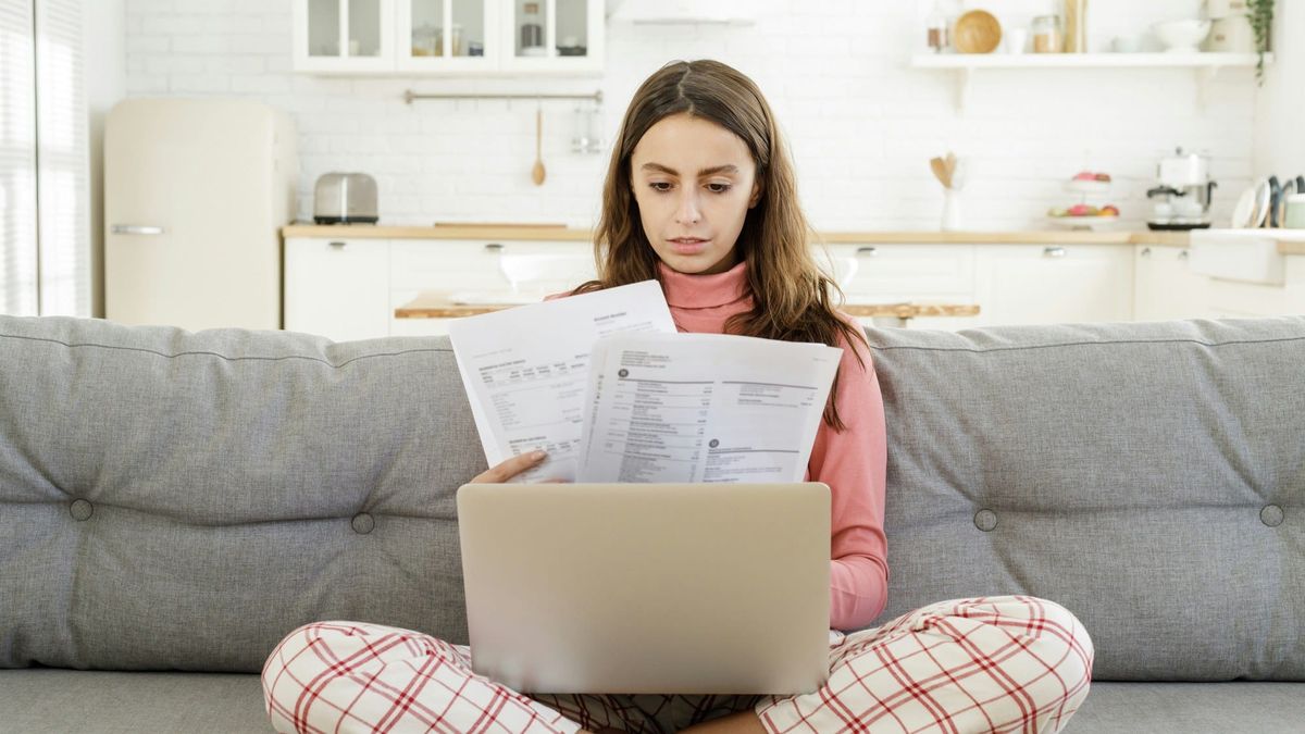 A woman sits cross-legged on a couch, reading through paper statements with a laptop open on her lap.