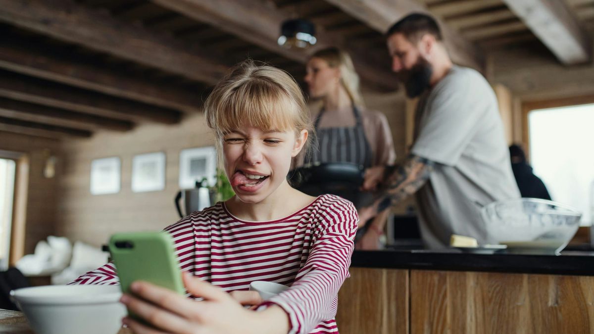 A girl looks at her phone at the kitchen table while her parents cook in the background.