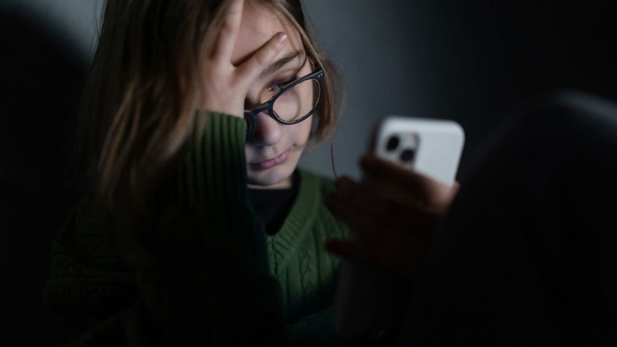 A young woman with one hand over her forehead, looking down at her phone in a dark room