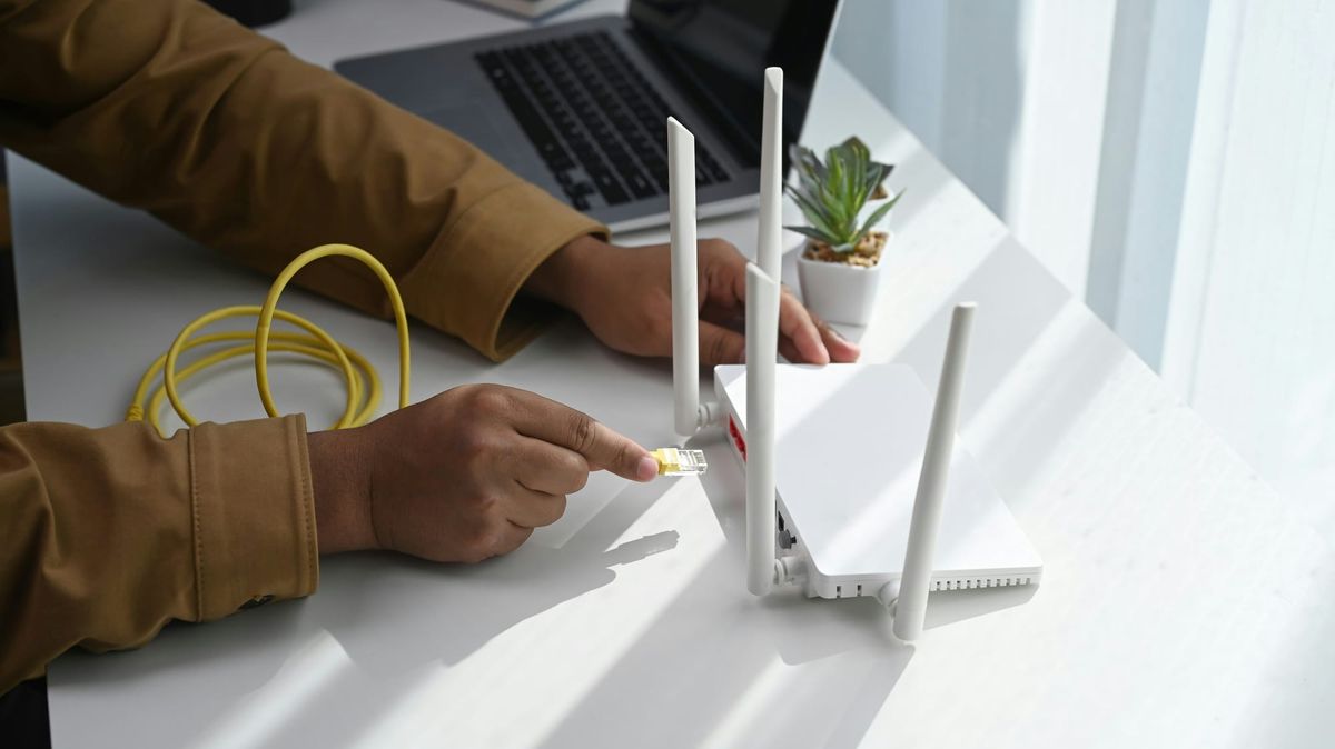 A person's hand unplugging the yellow network cable from the back of a white Wi-Fi router on a desk.