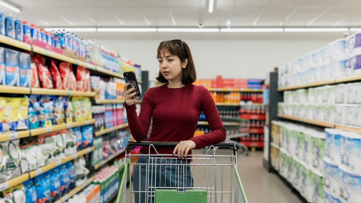 A shopper in a supermarket aisle looking at her phone, one hand on her cart.