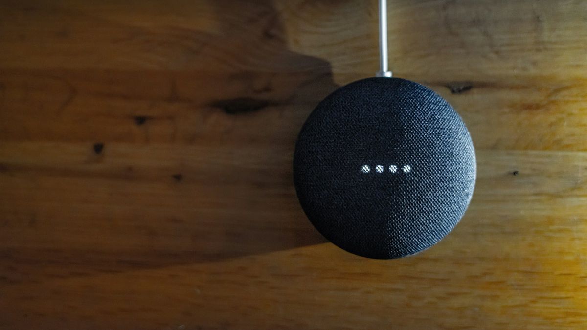 A charcoal Google Nest Mini smart speaker on a warm wooden wall, its four white status lights lit, with the power cable running in from above.