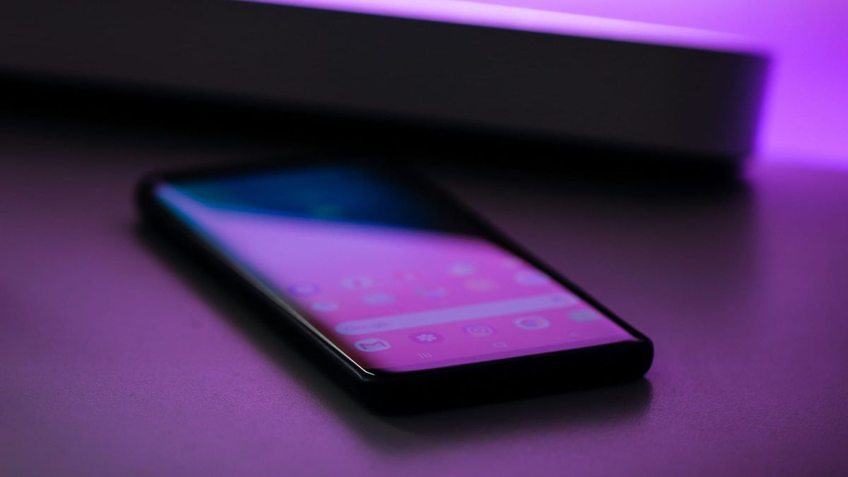A smartphone lying face up on a dark table with its home screen lit, a strip of purple light glowing on the wall behind it