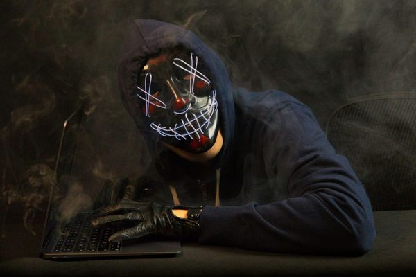 A hooded figure in a glowing stitched LED mask types on a laptop in the dark.
