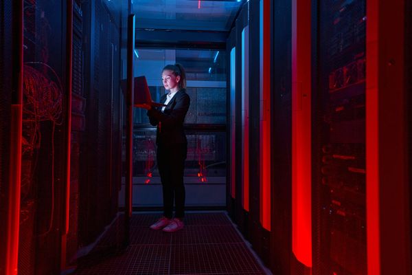 A security analyst works on a laptop in a dark, red-lit server room.