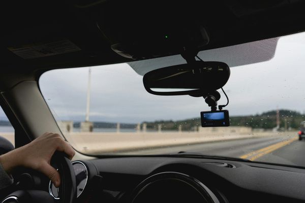 The view from a car's driver seat: a hand on the wheel, a small recording device by the mirror, an open bridge road ahead.