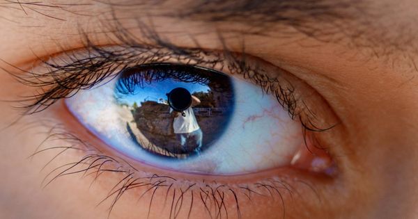 Extreme close-up of an eye, with a person holding a camera and a coastline reflected in the iris