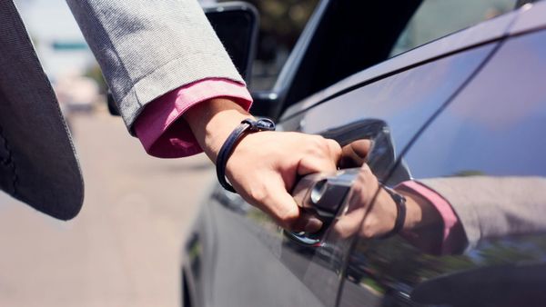 A hand pulling open the door handle of a dark sedan parked on a sunny street