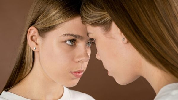 Two identical young women stand face to face, foreheads nearly touching, staring at each other
