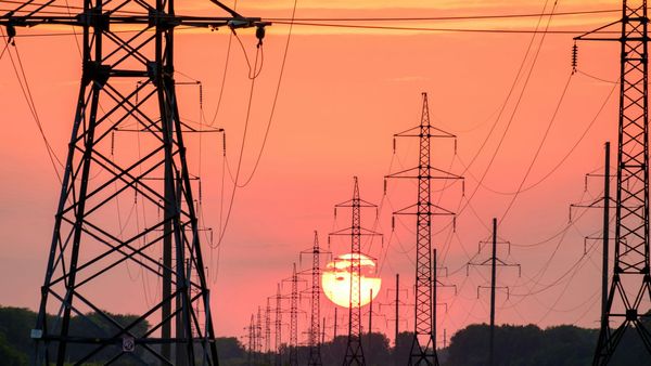 Rows of high-voltage power line towers silhouetted against a deep orange sky, with the sun low and huge behind them