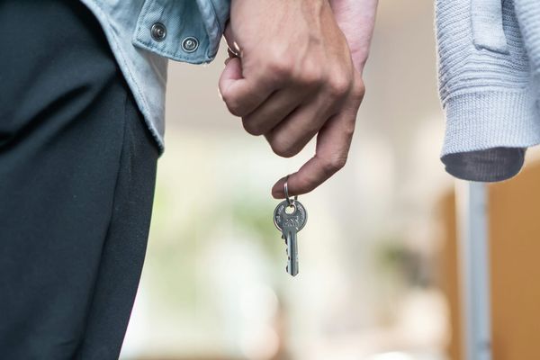 A hand holding a single house key on a ring, with a second person standing alongside.