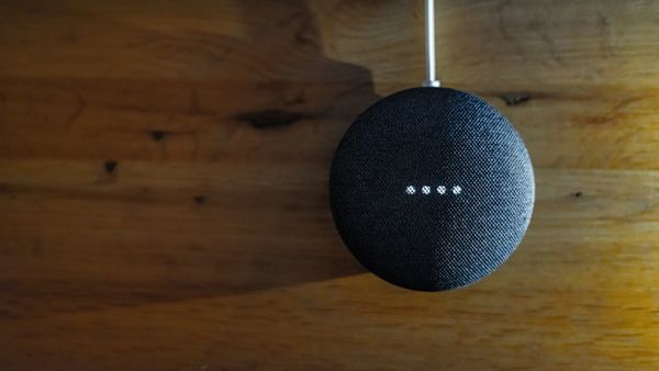 A charcoal Google Nest Mini smart speaker on a warm wooden wall, its four white status lights lit, with the power cable running in from above.