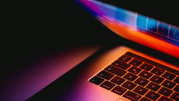 A MacBook nearly closed in the dark, with colorful light from its screen glowing through the gap