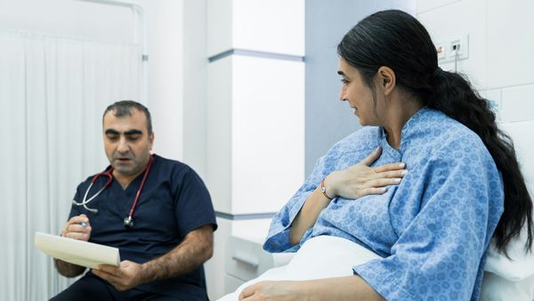 A doctor in navy scrubs sits with a notepad and pen while a patient in a hospital gown, her hand resting on her chest, describes her symptoms.