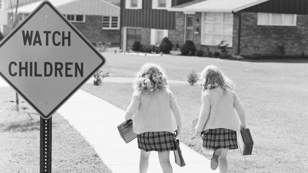 Two girls in school uniforms run up a sidewalk past a road sign reading WATCH CHILDREN