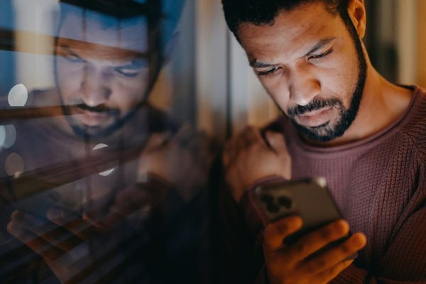 A man looks down at a phone in a dark room, his face repeated in the reflection of the window beside him.