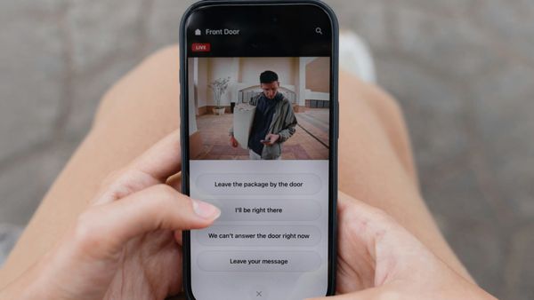 A phone held in both hands showing a video doorbell app, with a delivery driver holding a package on the live camera feed and quick-reply buttons below it.