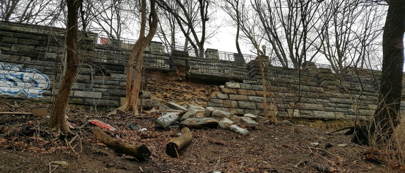 Chunks of lose stone sit below a broken retaining wall in a park.